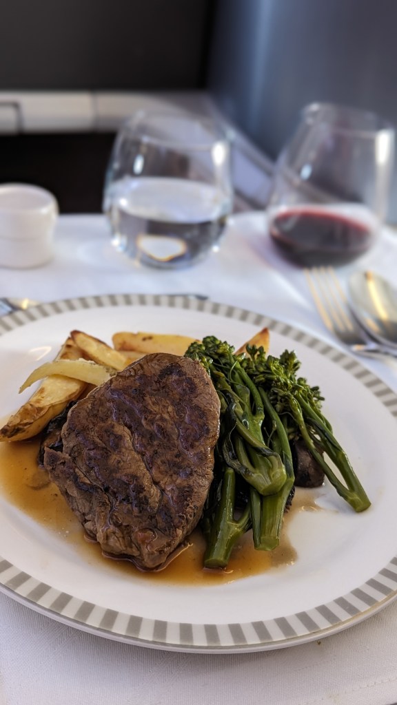 A plate featuring a grilled beef steak with green peppercorn sauce, accompanied by seasonal vegetables and roasted potatoes, set on a white tablecloth with glasses of water and red wine in the background.