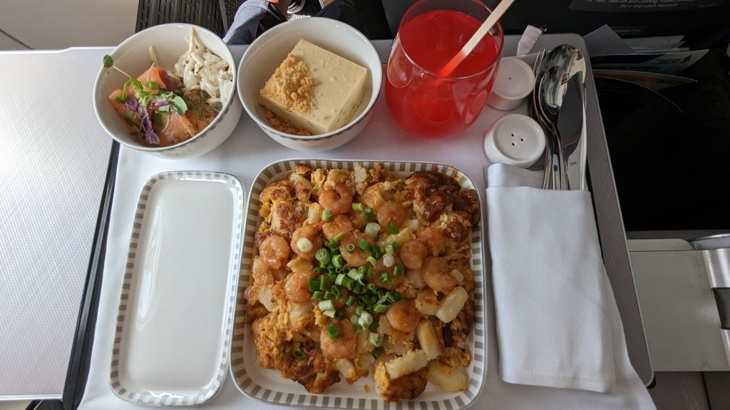An image of a Singapore Airlines meal tray featuring a serving of fried carrot cake topped with shrimp and green onions, accompanied by a bowl of salad with smoked salmon and a piece of dessert. A red drink is also present on the tray.