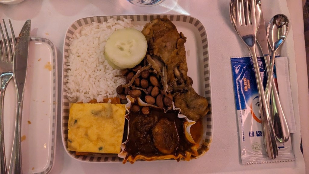 A served meal tray on an airplane featuring Nasi Lemak, consisting of fragrant rice, fried chicken, cucumber slices, peanuts, and a piece of omelette.
