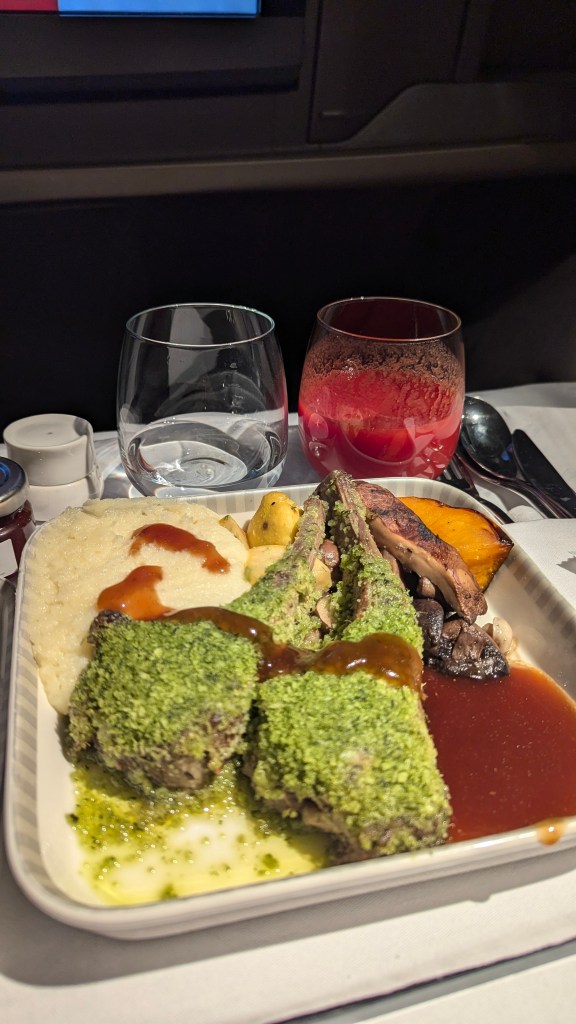 An airplane meal featuring two herb-crusted lamb chops on a tray, accompanied by mashed potatoes, roasted vegetables, and a sauce. Glasses of water and a red beverage are seen in the background.