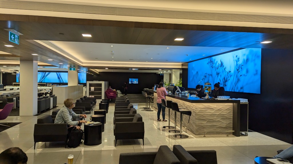 Interior view of a lounge with modern decor, featuring a bar area, seating arrangements, and a large screen displaying visuals. A few people are seen relaxing and enjoying refreshments.