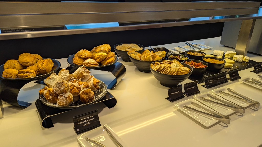 A display of assorted pastries and snacks in a lounge setting, including muffins, crackers, and various accompaniments, organized neatly on black trays.