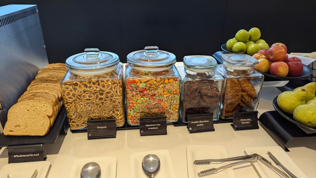 A display of snacks at an airport lounge, featuring jars of pretzels, Japanese rice crackers, double chocolate cookies, and cherry oat biscuits alongside a selection of wholemeal bread and fresh fruit including apples and pears.