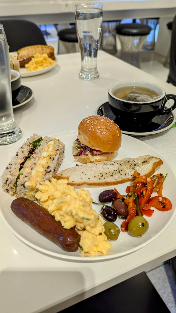 A plate of breakfast items featuring scrambled eggs, a sausage, a sandwich with visible fillings, an assortment of olives, red peppers, and a small sesame seed bun, accompanied by a black cup of tea and a glass of water.