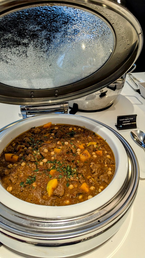 A close-up image of a covered serving dish containing lamb tagine with assorted vegetables, garnished with herbs, displayed on a dining table.