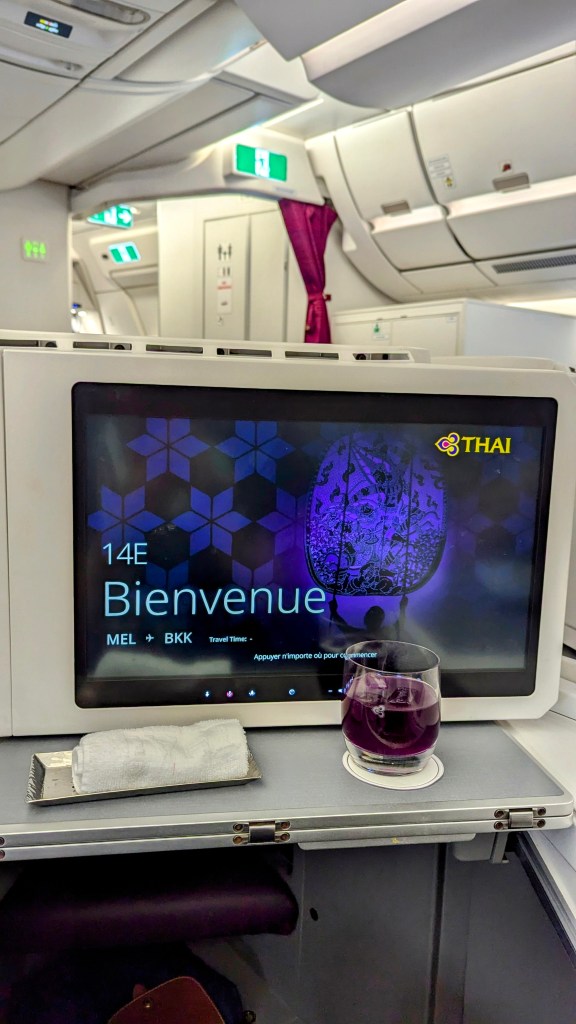 Interior view of Thai Airways 787-9 business class, featuring a personal screen displaying 'Bienvenue' and a purple drink in a glass.