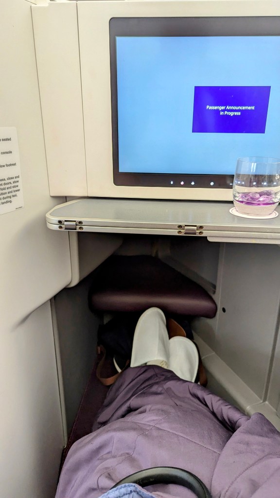 Interior view of a Thai Airways Boeing 787-9 business class seat with a passenger's feet resting on the footrest and a glass of water on the fold-out table. A screen displays a passenger announcement message.