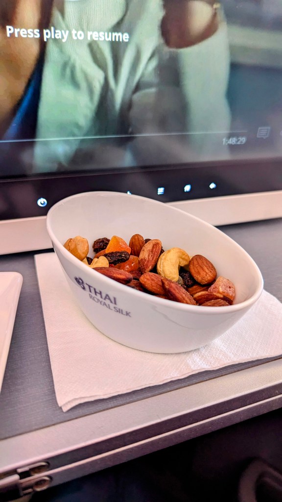 A bowl of mixed nuts served in a dish labeled 'Thai Royal Silk' on an airplane tray, with a screen displaying a paused video in the background.