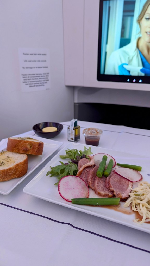 A beautifully presented plate of sliced meats and vegetables, accompanied by garlic bread and a small serving of sauce. A television screen can be seen in the background, displaying a scene from a program.