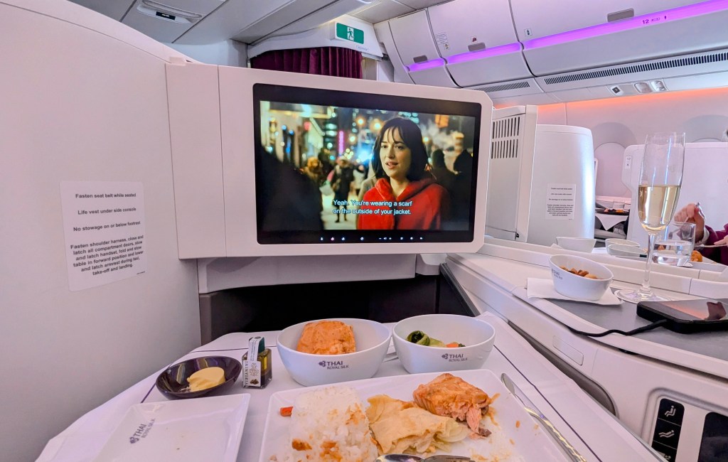 A view of a Thai Airways Boeing 787-9 business class seat with an entertainment screen showing a movie, a tray with leftover food, and a glass of champagne.