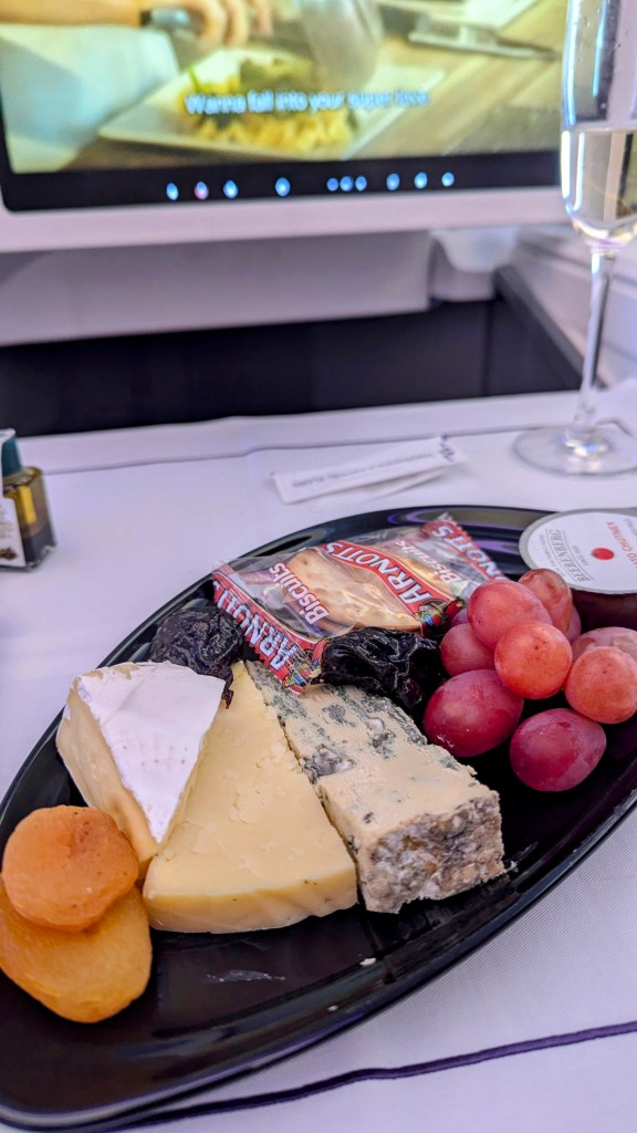 A close-up of a cheese platter in business class featuring various cheeses, dried fruits, and grapes, with a glass of champagne and an entertainment screen in the background.