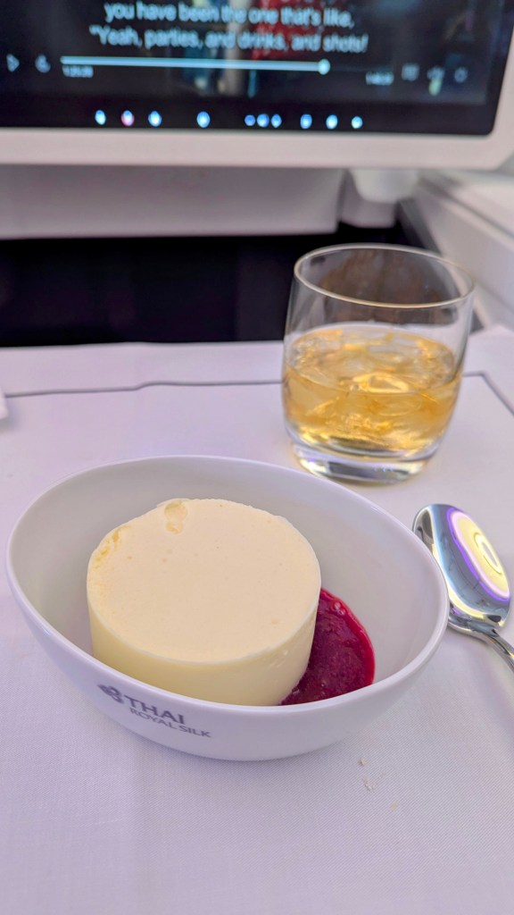 A dessert served in a white bowl with the Thai Airways logo, featuring a creamy mousse or panna cotta topped with berry sauce, accompanied by a glass of spirits, on a dining tray in a business class setting.