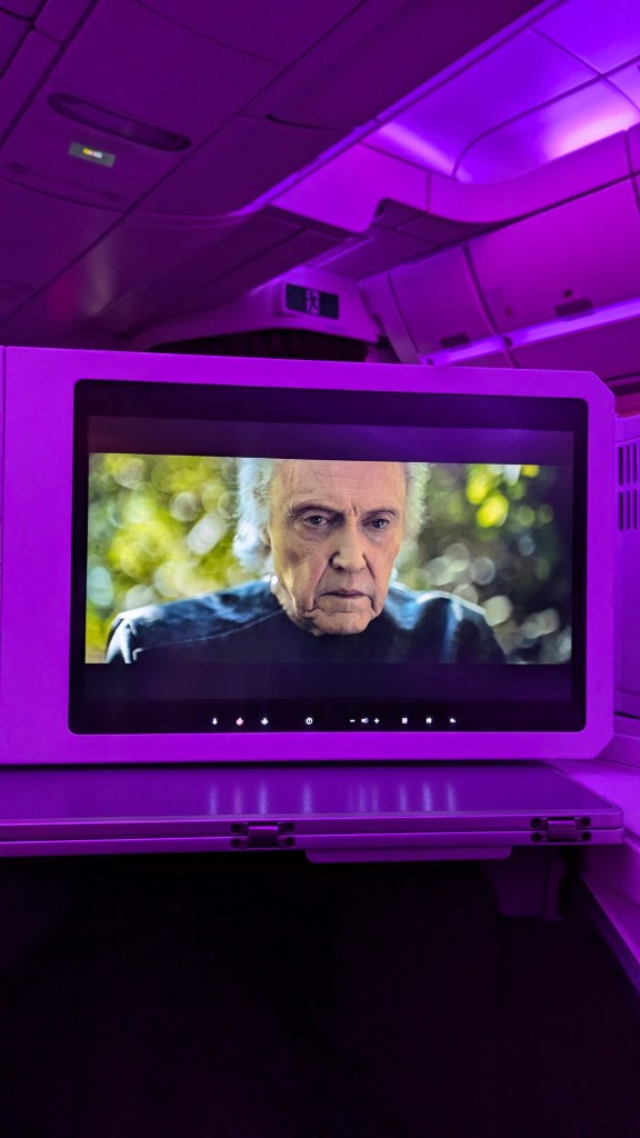 Close-up of a cabin in a Thai Airways Boeing 787-9 featuring a movie screen displaying a scene with a male character, surrounded by purple mood lighting.