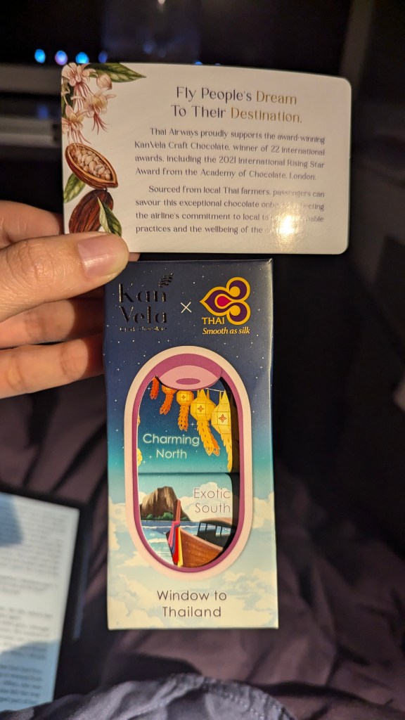 A hand holding a box of KanVela Craft Chocolate with colorful artwork showcasing Thailand's landscapes, accompanied by an informational card highlighting the chocolate's origin and Thai Airways' support for local farmers.